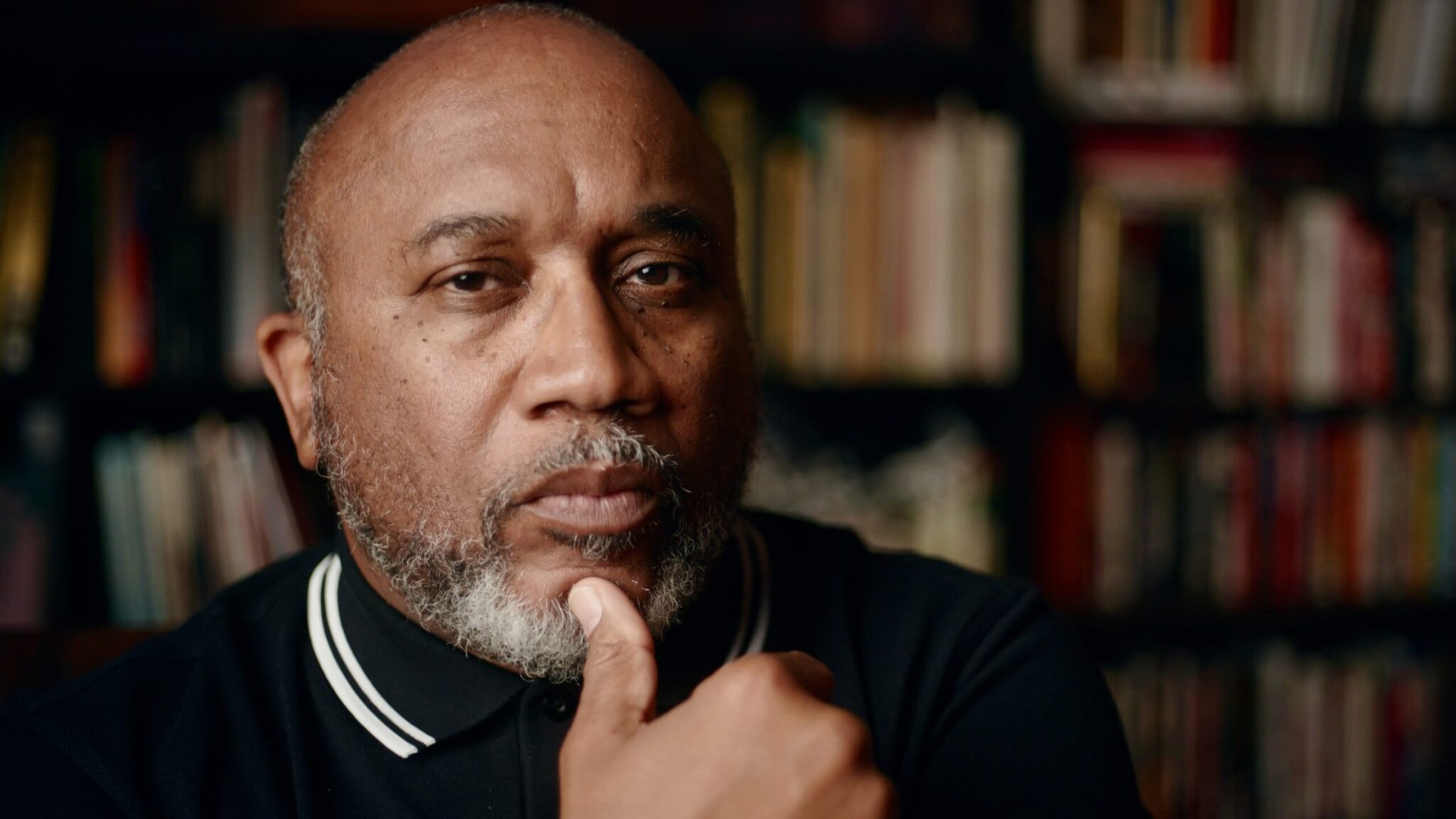 Photo of a black male looking directly into the camera. Gray and black beard. Black Fred Perry shirt with white tipping.